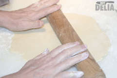 Pastry being rolled flat on a floured white food preparation board with a wooden rolling pin