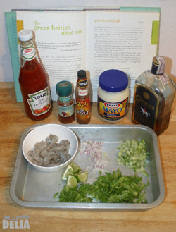 Aluminium tray containing fresh ingredients for prawn cocktail, with bottles and jars of other ingredients and a cookbook as a backdrop