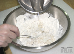 Water tipped into flour in a steel mixing bowl while a fork stirs the mixture