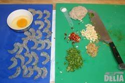 Cleaned prawns, an opened egg, a ball of bread and various chopped spices and herbs on a blue chopping-board for fish and a green chopping-board for vegetables