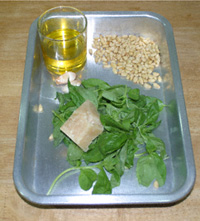 Basil, olive oil in a glass, pine nuts and Parmesan cheese, all in an aluminium roasting tin